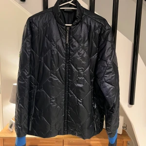 Svart quiltad bomberjacka J.Lindeberg - Quiltad svart bomberjacka från J.Lindeberg med snyggt mönster och diskret logopatch på ärmen. Jackan har dragkedja framtill, två sidofickor med dragkedja och blå muddar vid ärmslut. Materialet är glansigt och känns lätt, perfekt för dig som gillar stilren design.