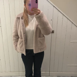 Beige teddyjacka från H&M Divided - Supermjuk och fluffig teddyjacka i beige från H&M Divided. Jackan har stora fickor framtill, där en av fickorna har ett litet hål, klassisk krage och stängs med knappar. Perfekt för dig som vill ha en mysig och trendig look under kyliga dagar.