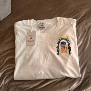 Vit t-shirt från Casablanca - Vit t-shirt från Casablanca med färgglatt tryck av byggnad och blad på både bröstet och ryggen. Klassisk rund hals och korta ärmar. Skön bomullskvalitet och snygg, avslappnad passform. Perfekt statement-plagg för dig som gillar unika prints.