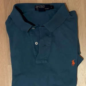 Mörkblå pikétröja från Polo Ralph Lauren - Snygg mörkblå pikétröja från Polo Ralph Lauren med klassisk krage och två knappar framtill. Tröjan har korta ärmar och den ikoniska orangea logon broderad på bröstet. Perfekt för dig som gillar stilrena och tidlösa plagg.