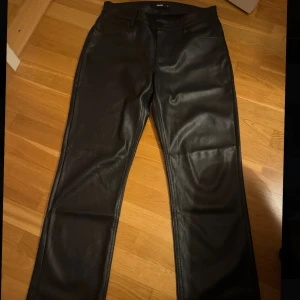 Svarta raka byxor i skinnimitation - Snygga svarta byxor från Bik Bok i skinnimitation med raka ben och klassisk femficksdesign. Perfekta för dig som vill ha en edgy look med stilren känsla. Byxorna har normal passform och stängs med dragkedja och knapp.