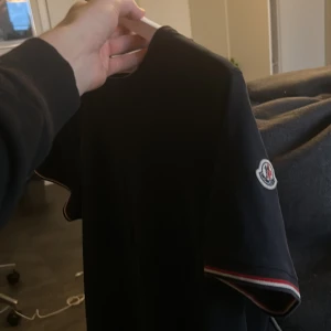 Navy blue Moncler T-Shirt med logga - Navy Blue t-shirt från Moncler med klassisk logga på ärmen. T-shirten har korta ärmar och smal passform. Ribbade muddar med röd och vit detalj vid ärmslut och halsringning. Tillverkad i mjuk bomull för en clean och stilren look. Passar till M & S