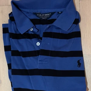 Blå och svart randig pikétröja Polo Golf - Snygg blå pikétröja från Polo Golf Ralph Lauren med svarta horisontella ränder och klassisk krage. Tröjan har korta ärmar, två knappar vid halsen och den ikoniska broderade loggan på bröstet. Perfekt för en chill och sportig look.