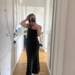 Svart långklänning med smala axelband - Säljer en svart långklänning med smala axelband och snyggt åtsittande passform. Klänningen har en enkel och stilren design med öppen rygg, perfekt för dig som gillar minimalistisk stil. Materialet känns mjukt och följsamt mot kroppen.
