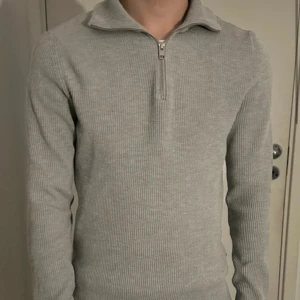 Grå ribbad half zip stickad tröja - Snygg grå stickad tröja med ribbad struktur och half zip-dragkedja framtill. Tröjan har en hög krage och långärmade ärmar, perfekt för dig som gillar stilren och enkel design. Passformen är normal och materialet känns mjukt och skönt mot huden.