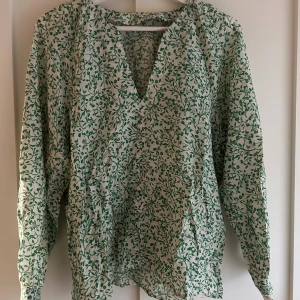 Grönblommig v-ringad skjorta Lindex  - Snygg v-ringad skjorta från Lindex i storlek S. Skjortan är vit med grönt blommönster, har långa ärmar och är tillverkad i mjuk bomull. Perfekt för dig som gillar en avslappnad men ändå trendig stil.