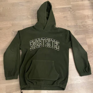 Grön hoodie från SWEET SKTBS - Mörkgrön hoodie från SWEET SKTBS med stor vit logga på bröstet. Tröjan har huva, känguruficka och ribbade muddar vid ärmslut. Perfekt för chill dagar och streetwear-stil.