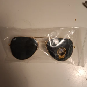 Ray-Ban aviator solglasögon guld - Snygga Ray-Ban aviator solglasögon med tunna guldfärgade metallbågar och mörka linser. Klassisk pilotform som ger en tidlös vibe och passar till de flesta outfits. Ray-Ban-logga syns på linsen. Perfekta för soliga dagar.