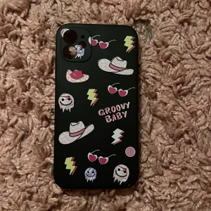 Svart mobilskal med motiv - Säljer ett svart mobilskal med färgglada motiv som cowboyhattar, hjärtformade solglasögon, blixtar och små spöken. Texten 'Groovy Baby' står på baksidan. Skalet är gjort av plast och har öppningar för kamera och knappar.