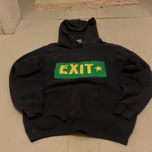 Blå hoodie EXIT Fruit of the Loom - Svart hoodie från Fruit of the Loom med stor EXIT-tryck i gult och grönt framtill och Ecole Française 99-tryck i vitt på ryggen. Klassisk huva, ribbade muddar och mjukt material i bomull och polyester. Perfekt för dig som gillar sportig och cool stil.