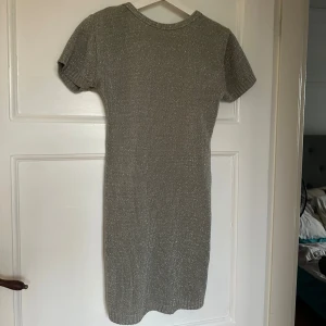 Glittrig silverklänning från QED London - Kortärmad, glittrig silverfärgad klänning från QED London med tight passform och djup, öppen rygg. Ribbad struktur och stretchigt material som ger en snygg siluett. Perfekt för dig som vill sticka ut med en cool look. Passar S