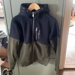 Svart-grön hoodie Sail Racing XS - Snygg hoodie från Sail Racing i storlek XS med mörkblått och olivgrönt färgblock. Tröjan har huva med snörning, hel dragkedja och en bröstficka med zip. Använd fåtal gånger och är i mycket bra skick. Perfekt för hösten och vintern, hör av er vid frågor!