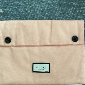 Ljusrosa liten necessär från Gucci Beauty - Säljer en söt liten ljusrosa necessär från G Beauty. Väskan har två svarta knappar framtill och är tillverkad i ett mjukt tygmaterial. Perfekt som accessoar för att bära småsaker. G-märket syns på en etikett på insidan.