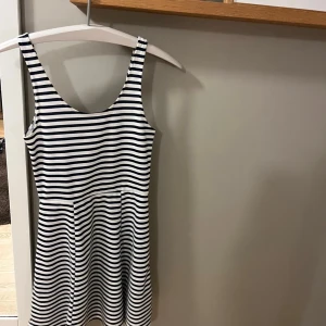 Randig kortklänning från Divided - Superfin kortklänning från Divided med marinblå och vita horisontella ränder. Klänningen är ärmlös med rundad urringning både fram och bak, och har en lätt utställd kjol. Perfekt för en fräsch och avslappnad look.