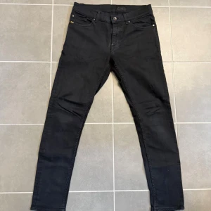 Svarta jeans från Tiger of Sweden - Svarta jeans från Tiger of Sweden, modell Evolve. Klassisk femficksdesign med smal passform och raka ben. Jeansen har diskreta nitar och logga vid myntfickan. Tillverkade i ett stretchigt bomullsmaterial för extra komfort.