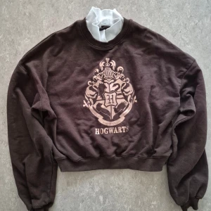 Tröja - Cool brun sweatshirt från H&M med Hogwarts-tryck i silver på bröstet. Tröjan har rund hals, långa ärmar och ribbade muddar vid ärmslut och nederkant. Materialet är mjuk bomull och passformen är normal. Perfekt för dig som älskar Harry Potter!