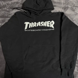 Svart Thrasher hoodie med tryck - Svart hoodie från Thrasher med stort vitt tryck på bröstet där det står 'Thrasher Skateboard Magazine'. Tröjan har huva med snörning och en klassisk magficka. Materialet är 90% bomull och 10% polyester, vilket gör den både mjuk och skön.