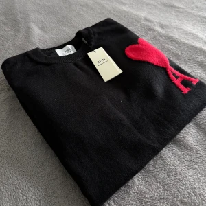 Ami Paris tröja  - Svart stickad tröja från Ami Alexandre Mattiussi med rund hals och ribbade muddar. På bröstet finns ett stort rött hjärta med bokstaven A, vilket ger en unik och snygg detalj. Perfekt för dig som gillar stilrena plagg med en twist.