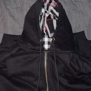 Burberry zip - Svart hoodiejacka från Burberry med klassiskt rutigt foder i huvan. Jackan har dragkedja framtill, snörning vid huvan och ribbade muddar. Snyggt broderad Burberry-logga på bröstet. Perfekt för dig som gillar stilren design med en twist.