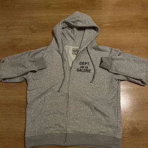 Grå hoodie från Gallery Dept. - Snygg grå hoodie från Gallery Dept. med svart tryck 'DÉPT. de la GALERIE' på både fram- och baksidan. Tröjan har huva, dragkedja och ribbade muddar. Tillverkad i mjuk bomull, perfekt för chill dagar. Storlek M.