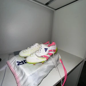 Adidas Predator fotbollsskor vit/rosa - Säljer ett par Adidas Predator fotbollsskor i vitt med rosa och neongula detaljer. Skorna har snörning, klassisk design och är tillverkade i syntetmaterial. Perfekta för dig som vill sticka ut på planen med både stil och funktion.