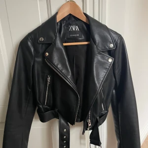 Svart skinnjacka från Zara - Snygg svart skinnjacka från Zara med croppad passform och coola detaljer som bälte i midjan och dragkedjor vid fickor och ärmslut. Jackan har klassisk krage med tryckknappar och ger en edgy vibe till din outfit.