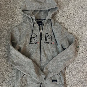 Grå hoodie från Race Marine med dragkedja - Säljer en grå hoodie från Race Marine i storlek S. Tröjan har huva med snörning, hel dragkedja och fickor framtill. På bröstet finns tryck med RM och koordinater. Mjuk insida och långärmad modell, perfekt för chill dagar.