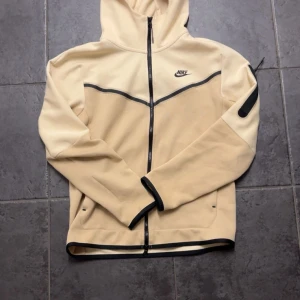 Beige Nike hoodie med svarta detaljer - Snygg beige hoodie från Nike med svarta detaljer och dragkedja hela vägen. Tröjan har huva, lång ärm och en praktisk ficka med dragkedja på ena ärmen. Tillverkad i en mjuk bomulls- och polyestermix. Perfekt för en sportig och avslappnad stil.