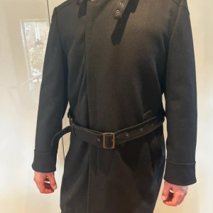 Svart kappa med bälte och dragkedja - Svart lång kappa med stilren design, bälte i midjan med metallspänne och dold dragkedja framtill. Jackan har lång ärm och diskreta sömmar på axlarna för extra detalj. 80% ull - perfekt för kyliga dagar och kvällar. 