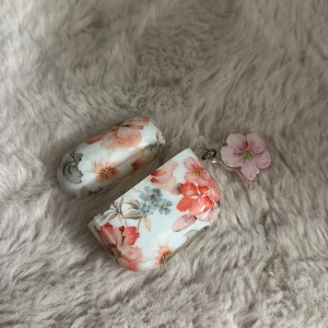 Blommigt fodral till airpods gen 3 - Snyggt blommigt fodral till trådlösa hörlurar, med rosa och blå blommor samt en dekorativ blomma som nyckelring. Fodralet är i gott skick med mindre slitage invändigt, perfekt för att skydda och ge din hörlurslåda en personlig touch.