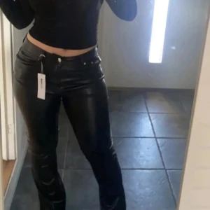 Svarta bootcut byxor i skinnimitation - Snygga svarta byxor med bootcut passform och hög midja i skinnimitation. Byxorna har klassisk femficksdesign, knapp och dragkedja framtill. Perfekta för dig som vill ha en edgy look med lite glans och attityd.