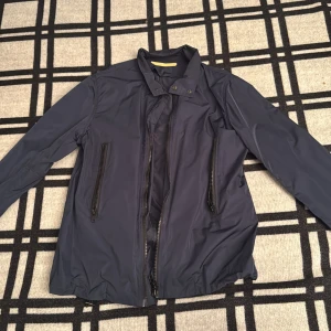 Mörkblå vindjacka från Whyred - Snygg mörkblå vindjacka från Whyred med hög krage och två stora dragkedjefickor framtill. Jackan har en stilren design med knappdetaljer vid halsen och är tillverkad i lätt syntetmaterial. Perfekt för dig som gillar minimalistisk och clean look.