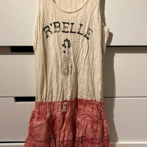 Rosa och beige klänning Scotch R'Belle - Snygg ärmlös klänning från Scotch R'Belle i beige och rosa. Överdelen har tryck med texten R'BELLE och en cool sjöjungfru, medan kjolen är volangig och rosa med virkade detaljer och knytband i midjan. Perfekt för dig som gillar en avslappnad och lekfull stil.