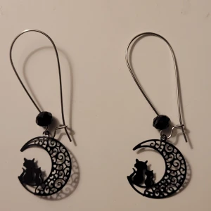 Svarta örhängen med katter och måne - Unika örhängen i svart metall med silvriga krokar. Hängsmycket har ett detaljerat mönster av en halvmåne med två katter som sitter tillsammans. Ovanför månen sitter en svart, facettslipad pärla som ger extra stil. Perfekta för dig som gillar djur och coola detaljer.