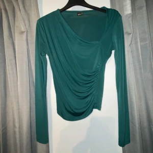 Grön omlottopp med drapering - Säljer en snygg grön omlottopp från ginatricot med draperad front och lång ärm. Toppen har en cool asymmetrisk v-ringning och är gjord i mjuk polyester som sitter skönt mot kroppen. Perfekt för dig som gillar stilrena detaljer och trendig look.