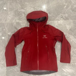 Röd Arc'teryx Gore-Tex vindjacka - Snygg röd vindjacka från Arc'teryx med Gore-Tex-material, perfekt för regniga dagar. Jackan har huva, hel dragkedja framtill och två praktiska fickor med dragkedja. Logga på bröstet och diskret Gore-Tex-märke på ärmen. Stilren och funktionell design.