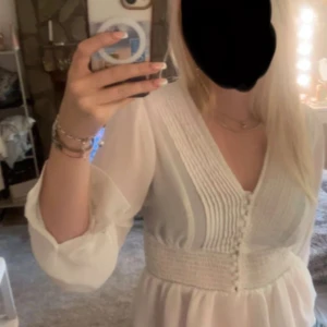 Vit blus med volang från Bik Bok - Supersöt vit blus från Bik Bok med v-ringning, smockad midja och volangkant nedtill. Blusen har långa, lätt transparenta ärmar och dekorativa knappar framtill. Perfekt för dig som gillar romantisk och feminin stil.