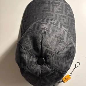 Svart Fendi keps med FF-mönster - Snygg svart keps från Fendi med det klassiska FF-logomönstret över hela kepsen. Kepsen har böjd skärm, justerbar rem bak och en diskret svart knapp på toppen. Materialet är slitstark polyester och kepsen har en lyxig vibe som sticker ut.