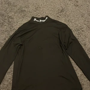 Svart polotröja med Los Angeles-tryck - Svart polotröja med lång ärm och coolt 'Los Angeles' tryck i vitt på kragen. Tröjan har en clean och slim passform, perfekt för dig som gillar streetstyle. Materialet känns mjukt och stretchigt, vilket gör den skön att bära hela dagen.