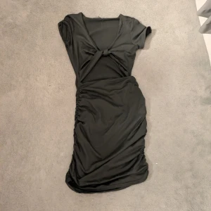 Svart kort klänning med knutdetalj - Svart kortärmad klänning med v-ringning och snygg knutdetalj framtill. Modellen är figurnära med rynkade sidor som ger en cool siluett. 