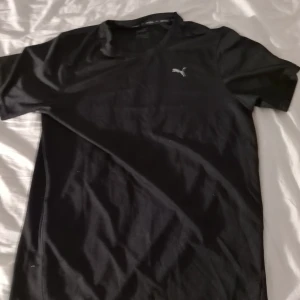 Svart t-shirt från Puma - Svart t-shirt från Puma med klassisk passform och korta ärmar. T-shirten har en liten vit Puma-logga på bröstet och är tillverkad i mjukt bomullsmaterial. Perfekt för en clean och sportig look.