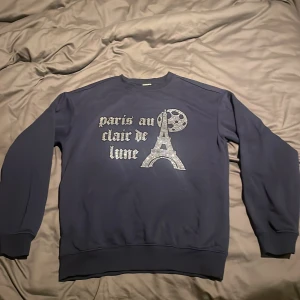 Mörkblå sweatshirt Paris Blinglucid -  mörkblå sweatshirt från Blinglucid med glittrigt tryck av Eiffeltornet och texten 'paris au clair de lune' i silver. Tröjan har rund hals, långa ärmar och ribbade muddar. Ny skick 🌟🌟
