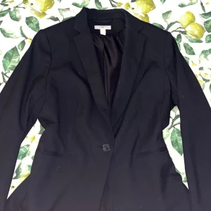 Svart kavaj från H&M, storlek 38 - Snygg svart kavaj från H&M i klassisk modell med en knapp framtill och två fickor. Kavajen har en stilren passform och är perfekt för dig som vill ha en enkel och tidlös look. Materialet är mjukt och känns bekvämt mot kroppen. Passar dig som gillar minimalistisk stil.