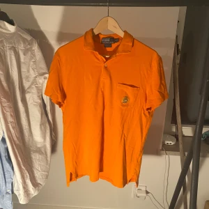 Orange pikétröja från Polo Ralph Lauren - Snygg orange pikétröja från Polo Ralph Lauren i bomull. Tröjan har klassisk krage, korta ärmar och en bröstficka med broderad detalj i guld, blått och rött. Custom fit-modell | Inga större defekter | 199 kr