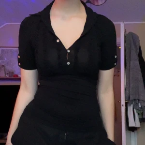 Svart kortärmad topp  - Svart figurnära topp från Intimissimi med korta ärmar och krage. Toppen har dekorativa knappar framtill och längs ärmarna, samt subtila volangdetaljer vid bröstet. Materialet är mjukt och stretchigt, perfekt för en clean och snygg look.