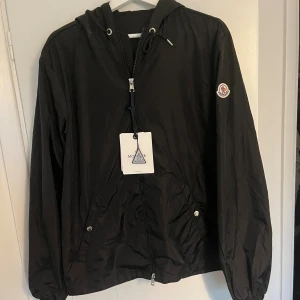 Svart vindjacka från Moncler med huva - Svart vindjacka från Moncler med huva och dragkedja framtill. Jackan har Moncler-logga på ärmen och snygga detaljer med snörning vid huvan samt fickor med knappar. Röd och vit rand på huvan ger extra stilpoäng. Perfekt för dig som gillar exklusiva streetwear-vibbar.