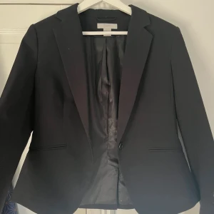 Svart kavaj från H&M - Stilren svart kavaj från H&M med klassisk krage och enkel knappstängning framtill. Kavajen har två fickor och är helfodrad med slät insida. Liten i storleken och är en kortare typ av kavaj 