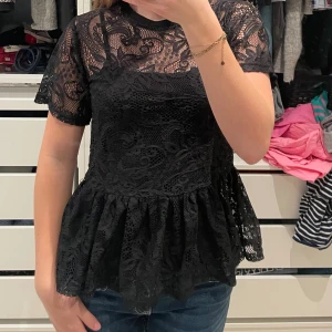 Svart spetsblus med volang - Supersnygg svart blus från Atmosphere i spets med blommigt mönster. Modellen har korta ärmar, rund halsringning och en volang nertill!  