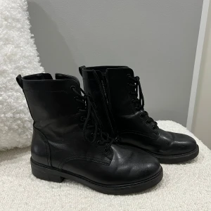 Svarta boots med snörning och dragkedja - Säljer ett par svarta boots i skinn med snörning framtill och dragkedja på sidan. Klassisk rund tå och platt sula, perfekt för dig som gillar en stilren och edgy look. Passar till både jeans och kjol för en cool vibe.