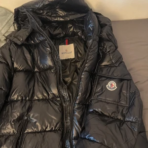 Svart Moncler maya jacka S - Svart pufferjacka från Moncler i glansigt material med huva och dragkedja. Jackan har en ficka med Moncler-logga på ärmen och quiltad design. Perfekt för dig som vill ha en snygg och varm jacka med street vibe.